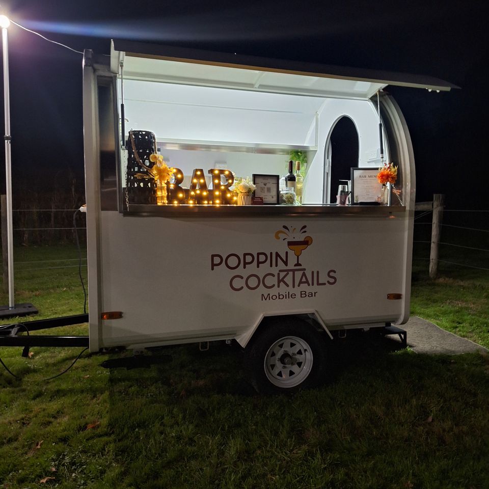 Poppin Cocktails mobile bar trailer illuminated at night with bar signage, decor, and beverage service setup for an event.