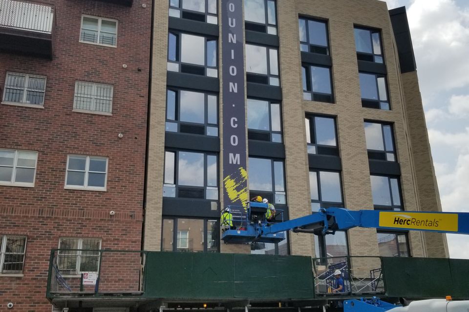Tall vertical printed banner running six storeys up a new building, installed from a boom lift