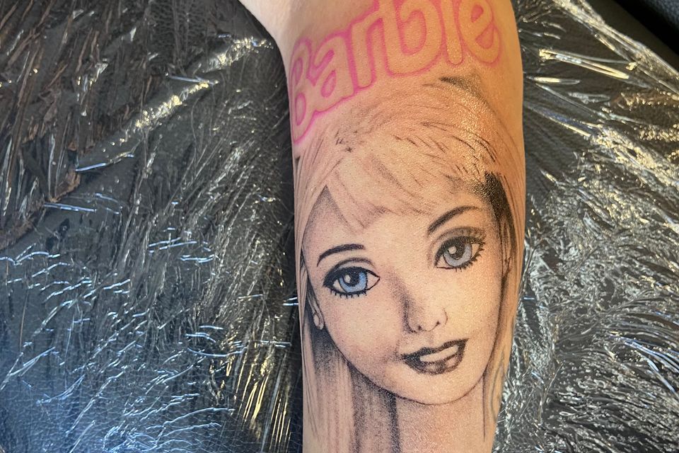 Image of a womans left forearm tattoo of Barbie in Black, Grey, Blue eyes, and red with white highlights