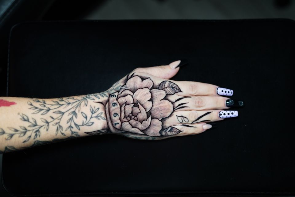 Image of a hand tattoo in great mostly black and white detail. Some green color for the branches and leaves on the forearm and the number 1993 in green color as well. Has 2 leaves and the letter J and what appear to be more sprigs of plants