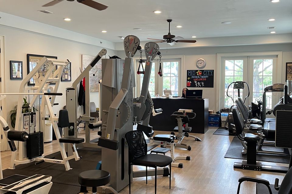 A spacious physical therapy gym at Mid City Physical Therapy in New Orleans, equipped with various exercise machines including a V1R leg press, treatment tables, a ceiling fan, and a large mirror along the wall, with a plant and water cooler in the corner