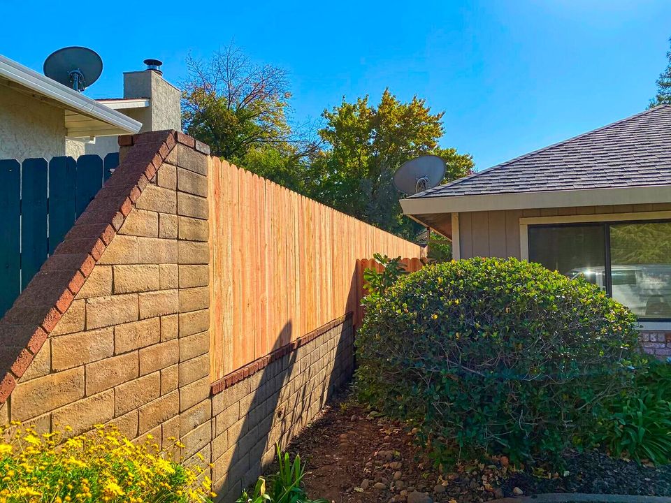 Enhanced view of a residential backyard with a new wood privacy fence atop a concrete retaining wall, bordered by yellow flowers and manicured shrubs, with neighboring homes and trees under a clear blue sky