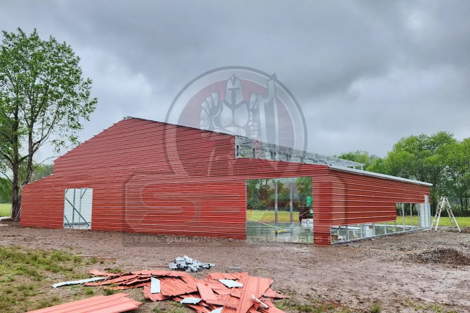Red steel barn under construction with open bays and framing with SBSI watermark representing steel barns in Alabama