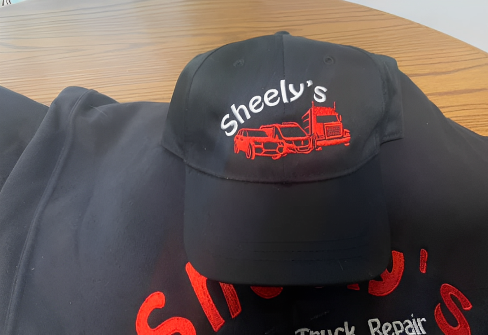 Black embroidered cap featuring a custom Sheely's Truck Repair logo in white and red stitching.
