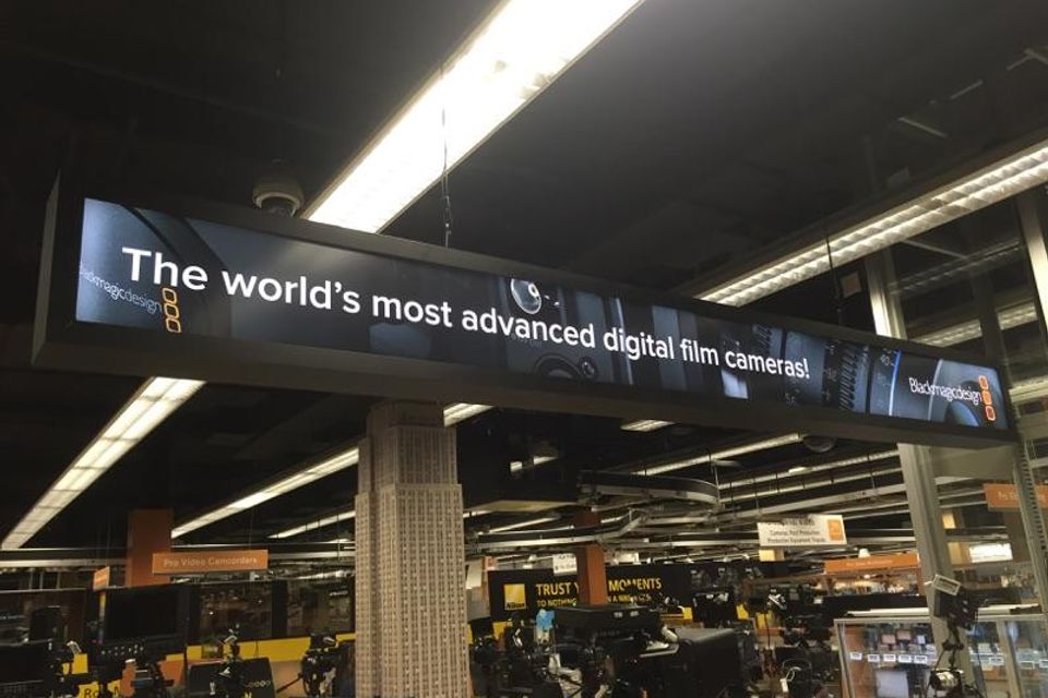 Long printed overhead sign panel suspended from the ceiling above a camera store sales floor