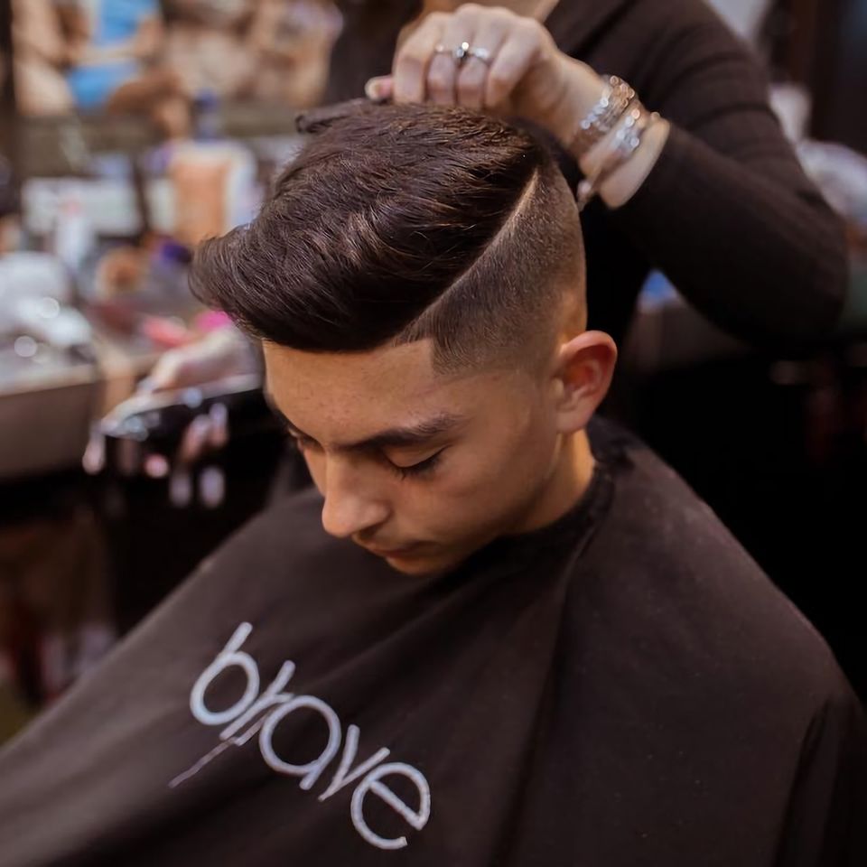 Close-up of a barber cutting a client’s hair with scissors and a comb, while a counter displays a hairdryer, spray bottles, and other grooming essentials.