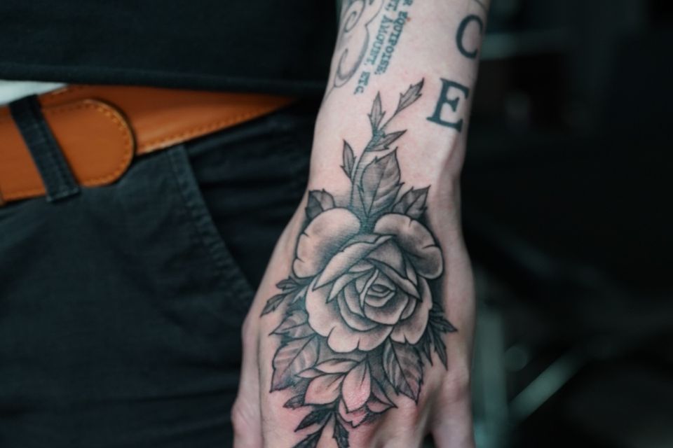 Image of a rose tattoo on a man's left hand with additional foiliage of leaves in Black & Grey