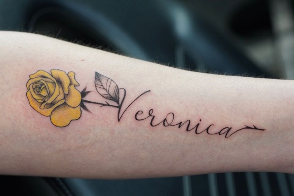 Image of a woman's left arm with the name Veronica tattood in cursive and a yellow flower with black outlines and a black stem to it's left