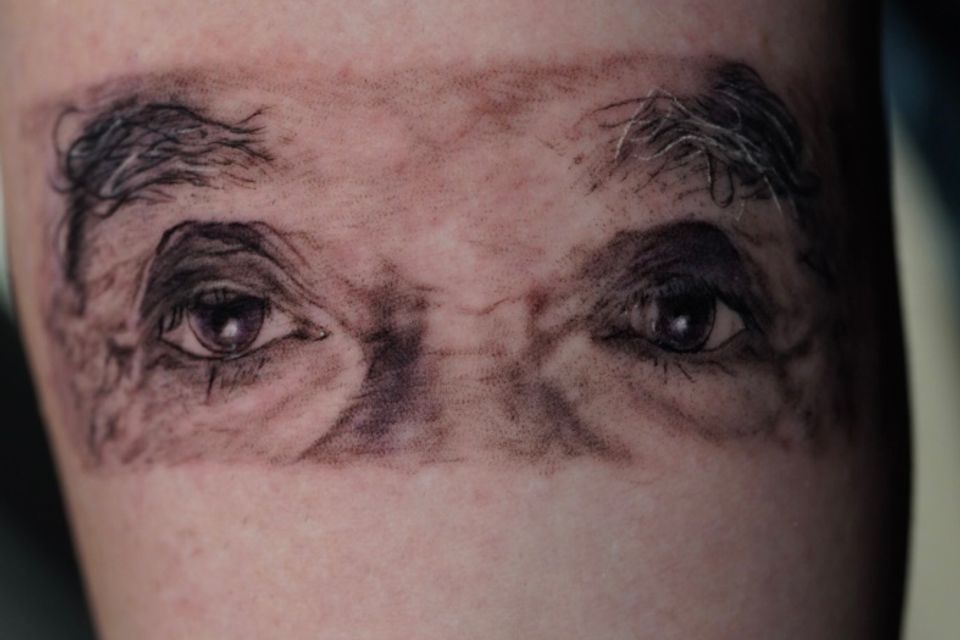 Image of a black and grey tattoo of eyes, eyebrows and part of the bridge of the nose framed in a rectangular shape in the crook of the right elbow