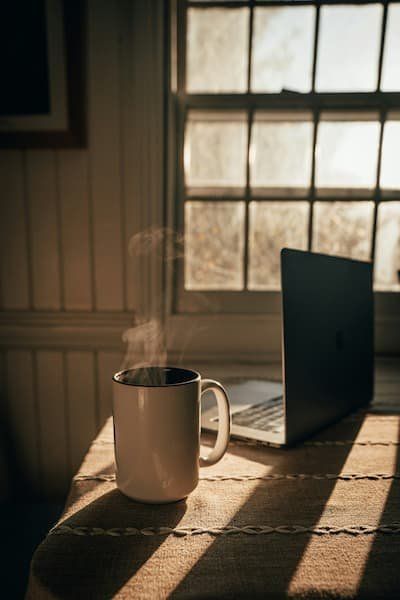 Cup of steaming coffee by open laptop with sun shining through the window 