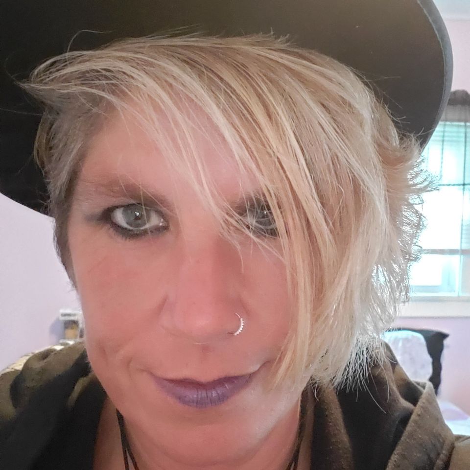 Close-up portrait of Kelly wearing a black hat and silver nose ring, smiling confidently.