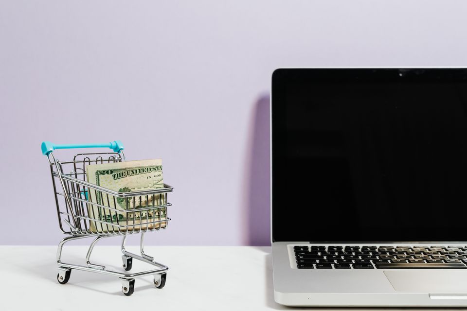 Laptop next to a miniature shopping cart with money inside it. Concept of e-commerce.