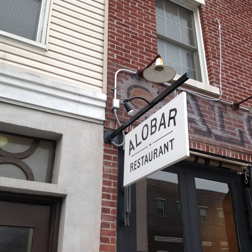 White rectangular blade sign on a black bracket with gooseneck lights above a restaurant entrance