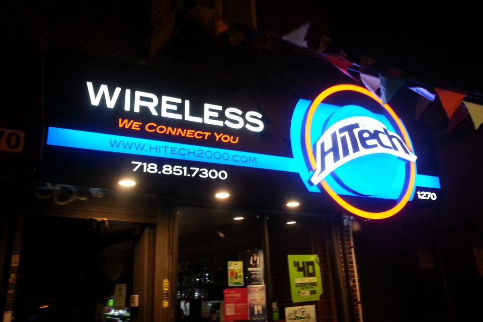 Illuminated HiTech Wireless sign lit at night, with a glowing blue and orange circular logo and white lettering above a storefront