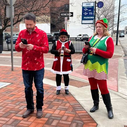 Three attendees dressed in Christmas-themed outfits standing on a downtown sidewalk while looking at their phones.