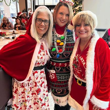 Three women wearing Christmas-themed clothing and accessories smiling for a photo indoors.