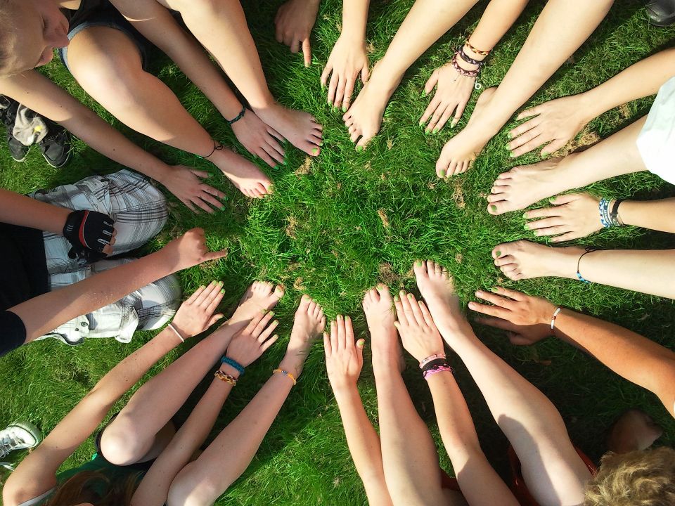 A group of people sitting in a circle on grass, with their hands and feet extended towards the center, creating a sense of unity and teamwork.