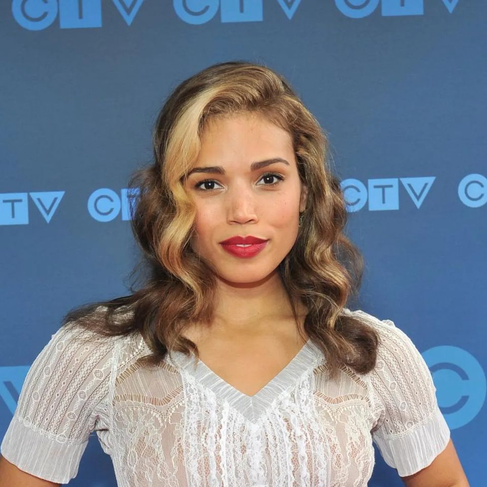 Actor Ciara Renée posing in front of a blue CTV backdrop, wearing a white blouse with wavy shoulder-length hair and red lipstick, appearing at a media event.