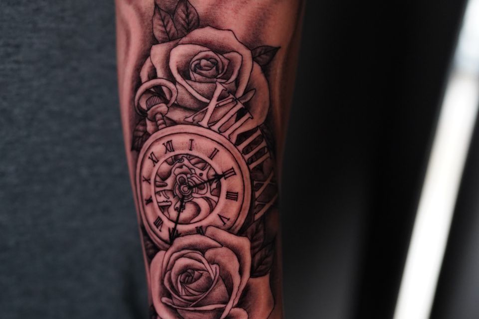 Image of a man's forearm with a very detailed tattoo with the name Bertha above the detailed Roses, Clock and background in black and grey ink