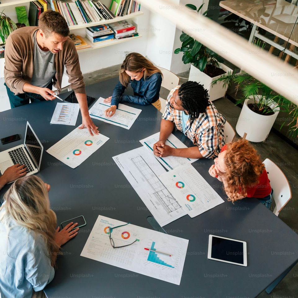 Group of six people sitting at a table in a modern office, surrounded by charts, laptops, and documents, engaging in discussion.
