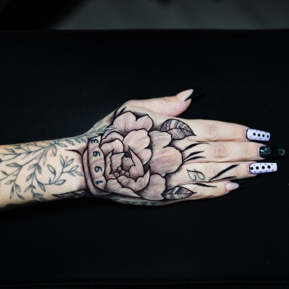 Image of a hand tattoo in great mostly black and white detail. Some green color for the branches and leaves on the forearm and the number 1993 in green color as well. Has 2 leaves and the letter J and what appear to be more sprigs of plants