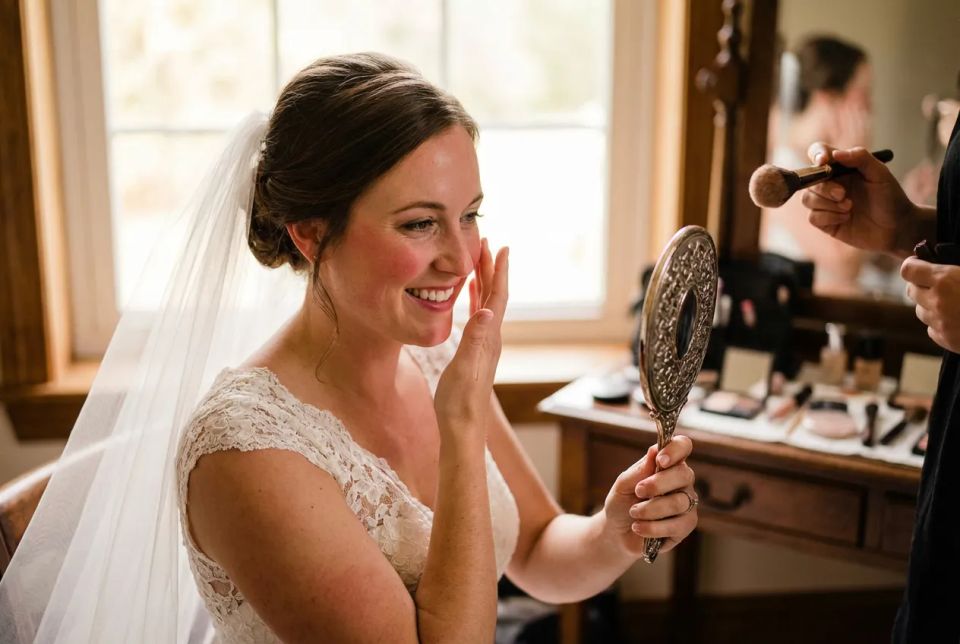 Smiling bride looking in a vintage handheld mirror while a makeup artist applies finishing touches before the wedding ceremony