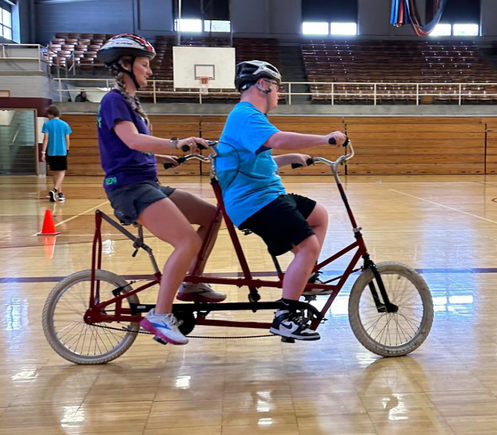 A participant and volunteer ride a tandem bicycle together inside a gymnasium during DSAV's iCan Shine Bike Camp.