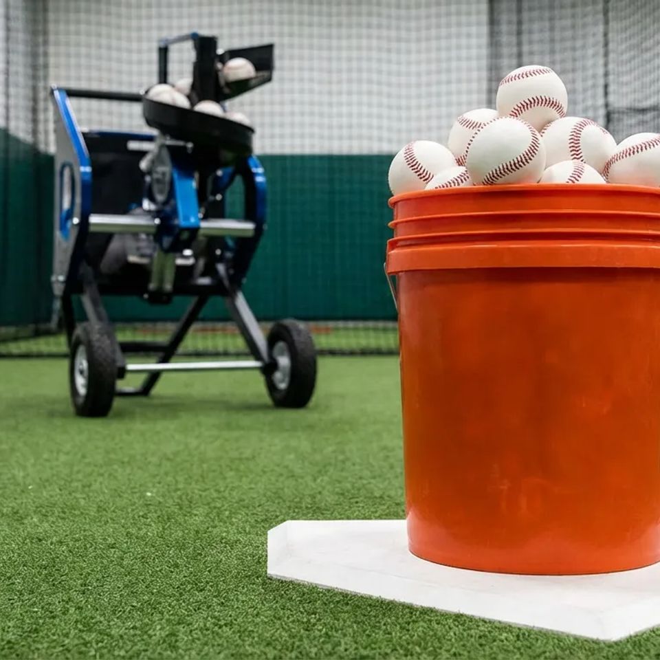 Orange bucket full of baseballs sitting on home plate inside an indoor batting cage with an automatic pitching machine in the background.