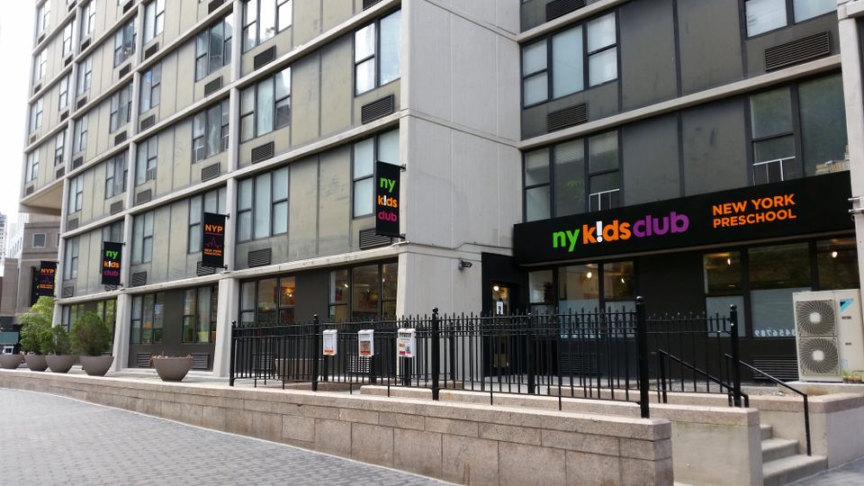 Row of NY Kids Club flag signs projecting along a building facade above a New York Preschool storefront sign in Battery Park