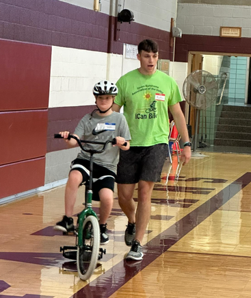 A volunteer coach walks beside a child riding an adaptive bicycle during a one-on-one training session at DSAV's iCan Shine Bike Camp.