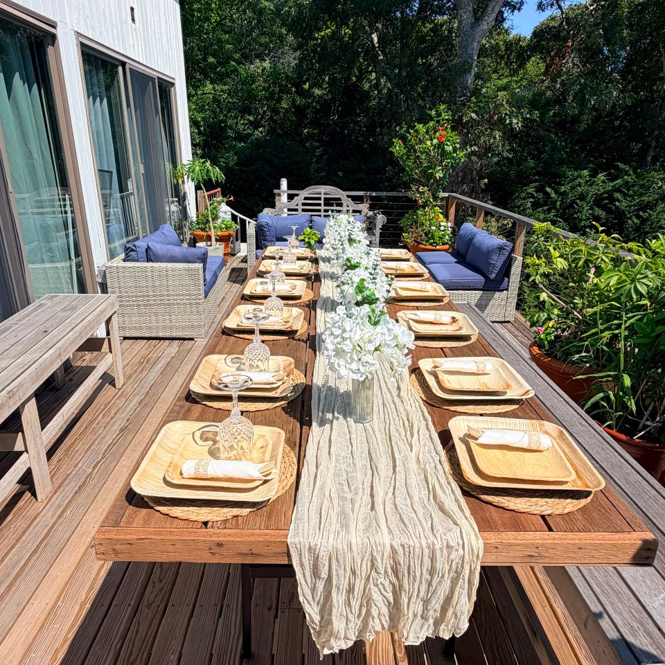 Long outdoor dining table styled with palm plates and floral runner on a deck for a private event in the Hamptons NY