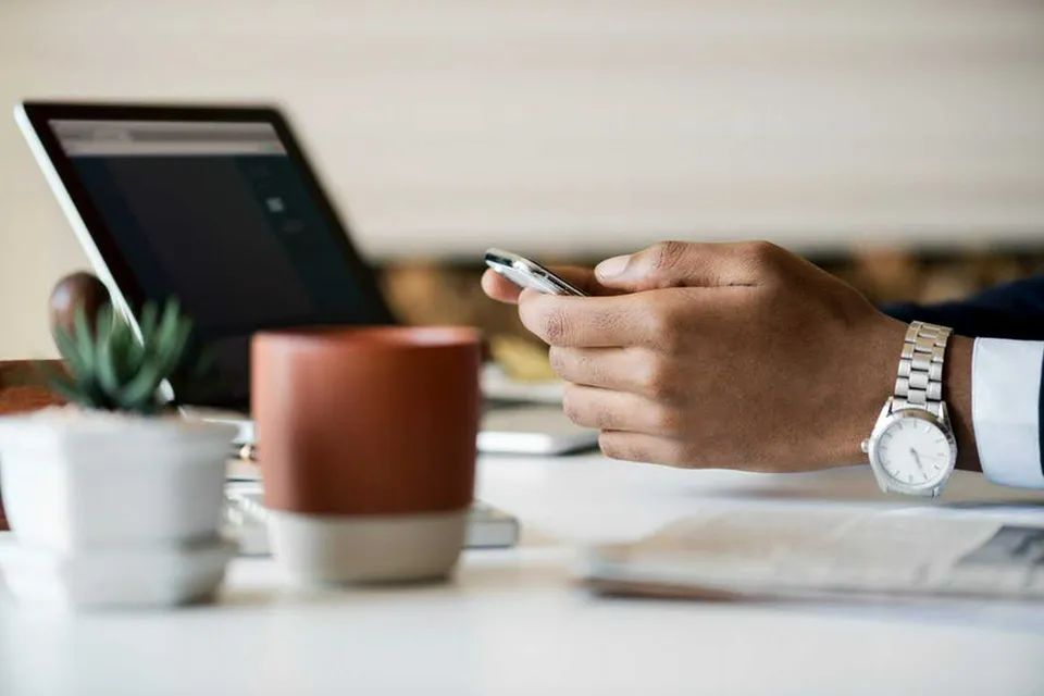 Man wearing a watch holds his phone in front of him. Laptop in the background. Only the man's hands and wrists are visible.