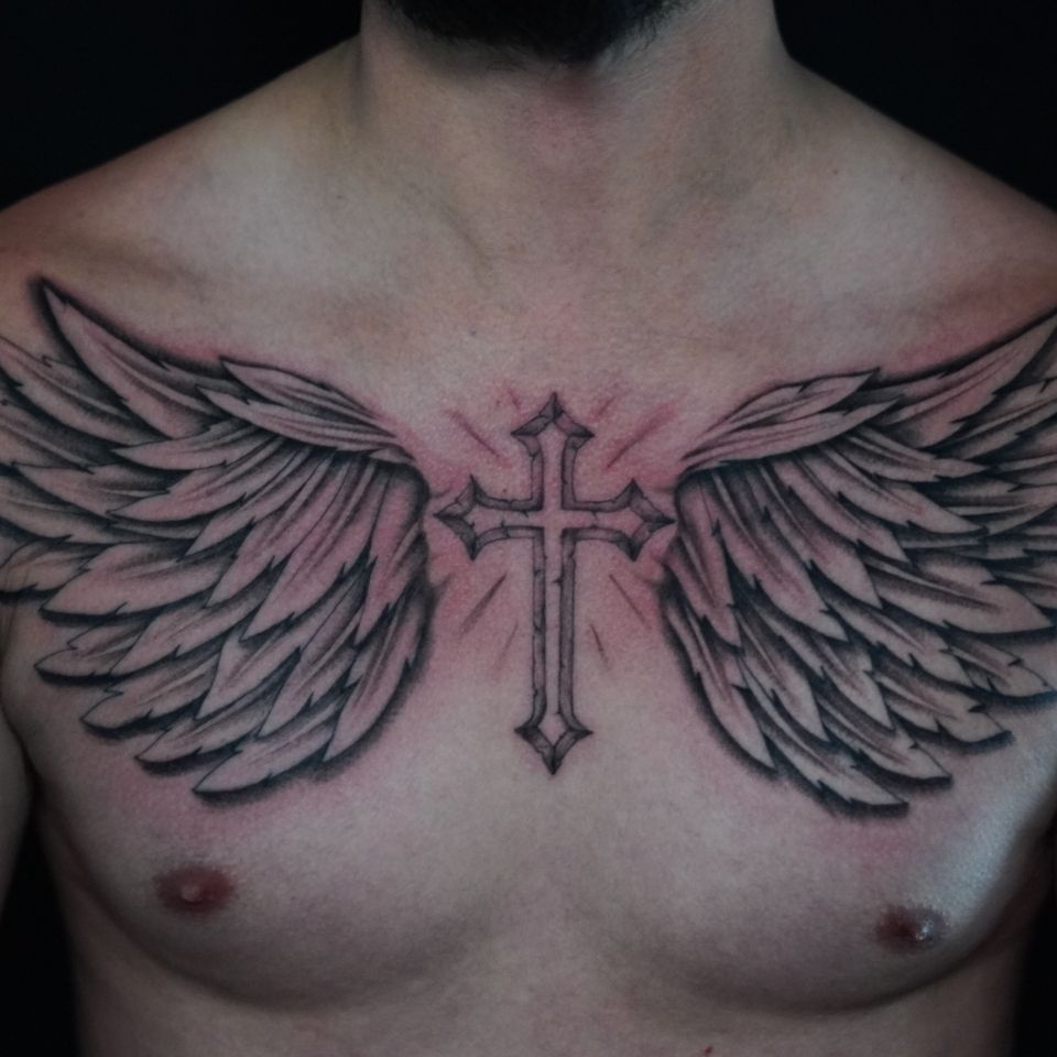 Image of a pair of wings tattood on a mans chest with a cross in the center between the wints