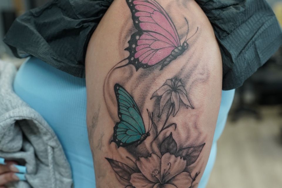 Image of a tattoo that includes 2 colorful butterflies, 1 pink and black and one teal and black, while the garden of flowers is in black and shades of grey