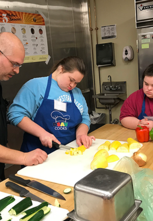 Participant with Down syndrome practicing knife skills under instructor supervision during a cooking class.