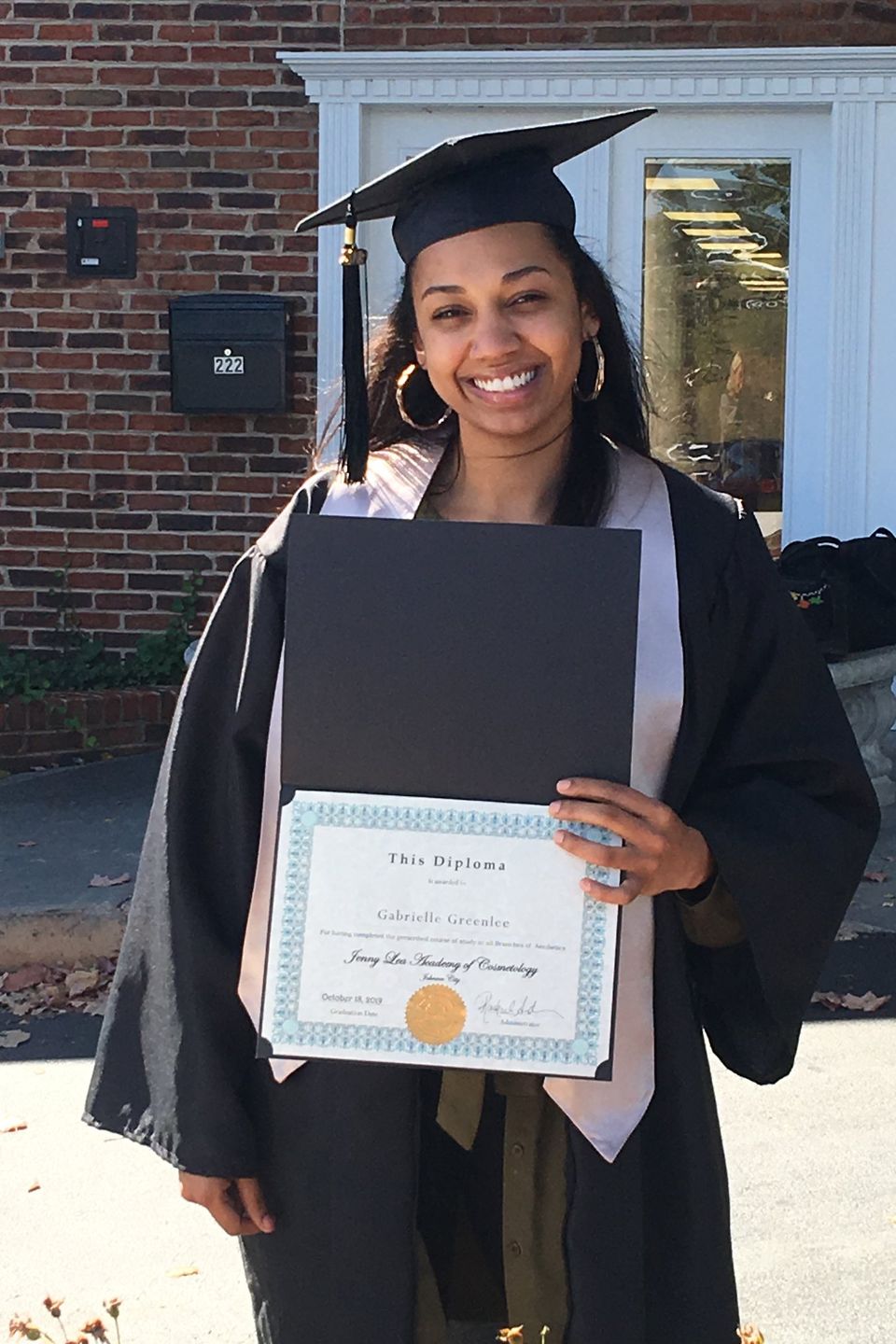 Cosmetology graduate holding diploma outside Jenny Lea Academy