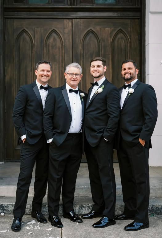 Groom, groomsmen, and father of the groom wearing matching black tuxedos from New Orleans Tuxedo Inc., posing for wedding photos outside a church.