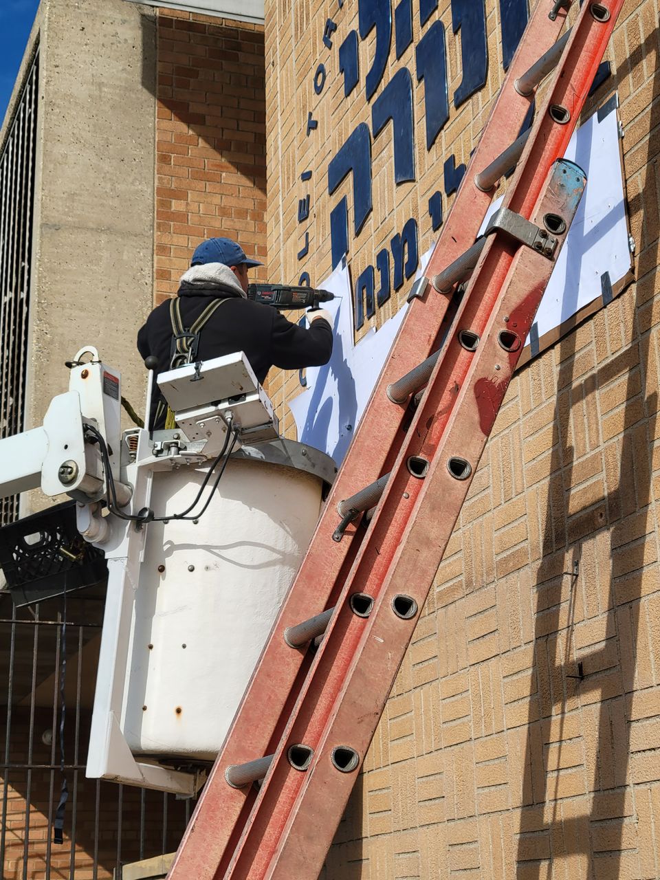 Installer harnessed in a bucket lift drilling a sign panel into a brick facade