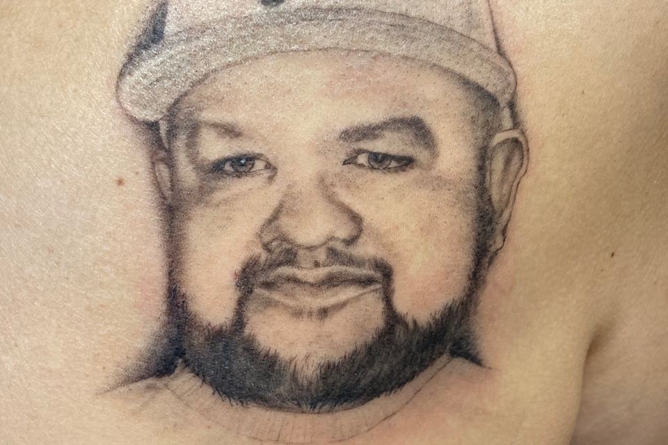 Image of a tattoo of a man with a beard in a Pirates ball cap on a womans left back shoulder in black and grey