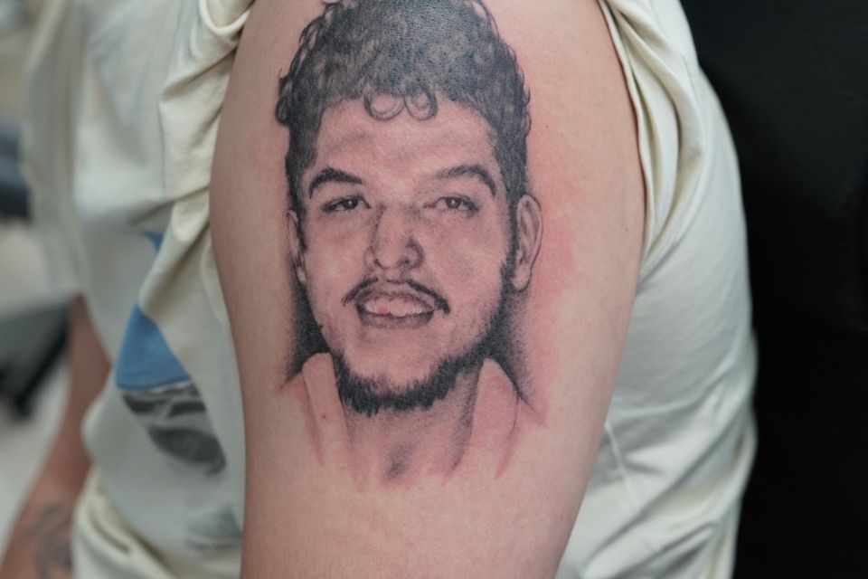 Image of a man with a tattoo on his left arm of a man with curly hair, pronounced eyebrows, pencil thin mustache and scruffy beard in black and grey ink