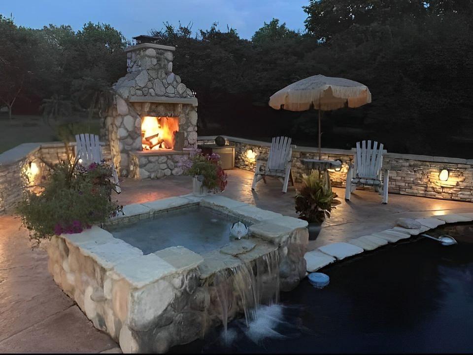 Stone outdoor patio featuring a pool, fireplace, seating area, and landscaped surroundings.