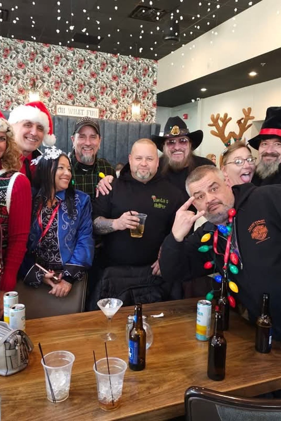 Group of attendees wearing Christmas-themed clothing and accessories gathered around a table at a holiday party indoors.