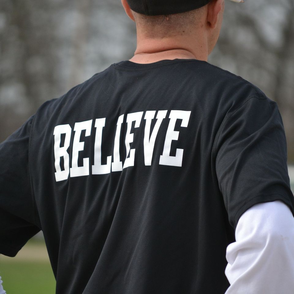Athlete wearing a black performance shirt with the word “BELIEVE” printed in large white letters across the back.