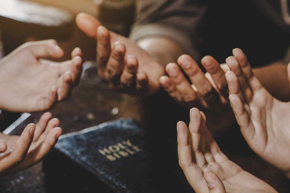 Several people gathered with hands raised in prayer around an open Holy Bible during a worship and fellowship gathering.