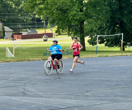 A participant rides an adaptive bicycle outdoors while a volunteer coach runs alongside providing support during DSAV's iCan Shine Bike Camp.