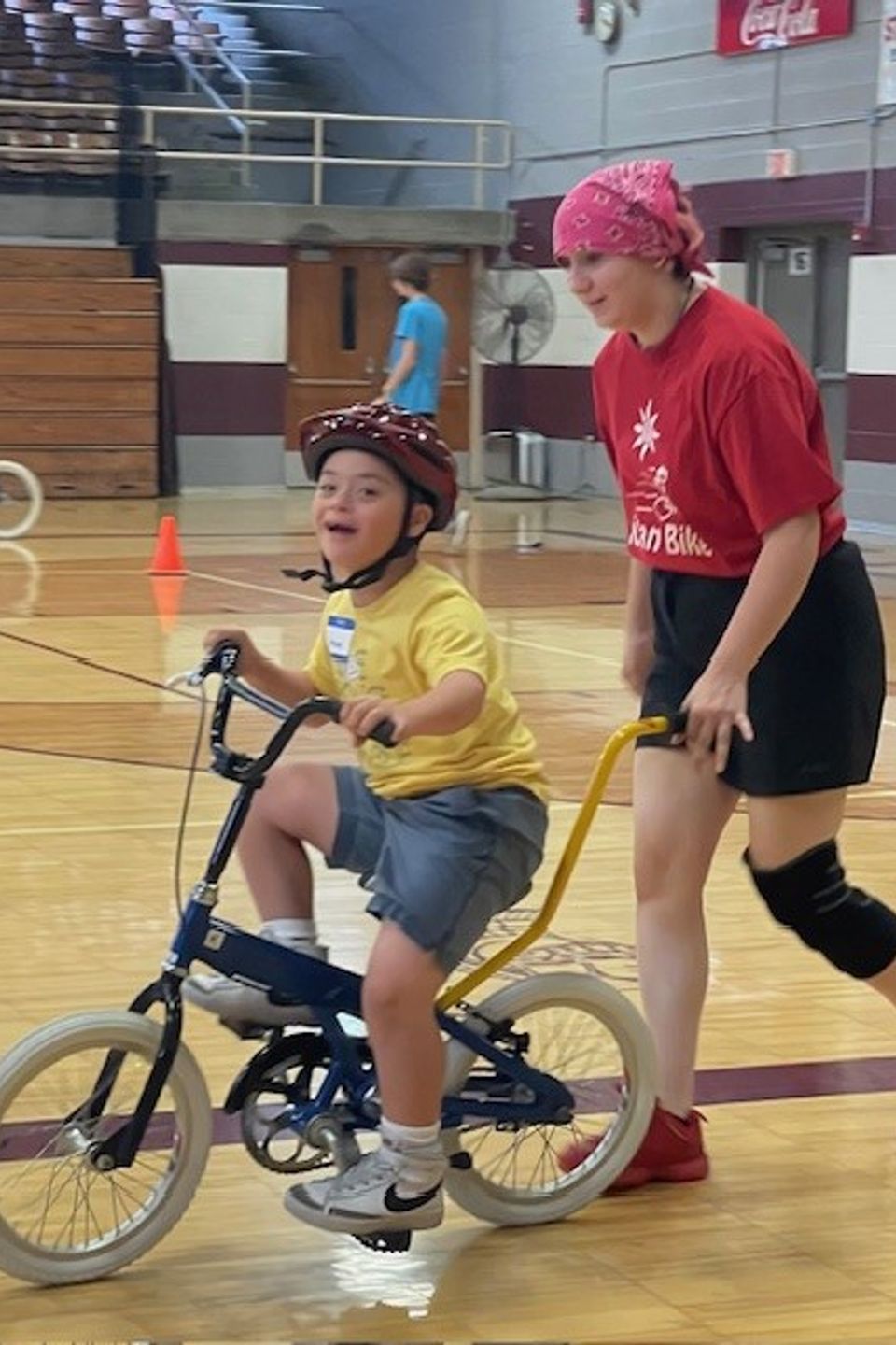 Volunteer coach assisting a child with Down syndrome while riding an adaptive bicycle during the DSAV iCan Shine Bike Camp in a gymnasium.