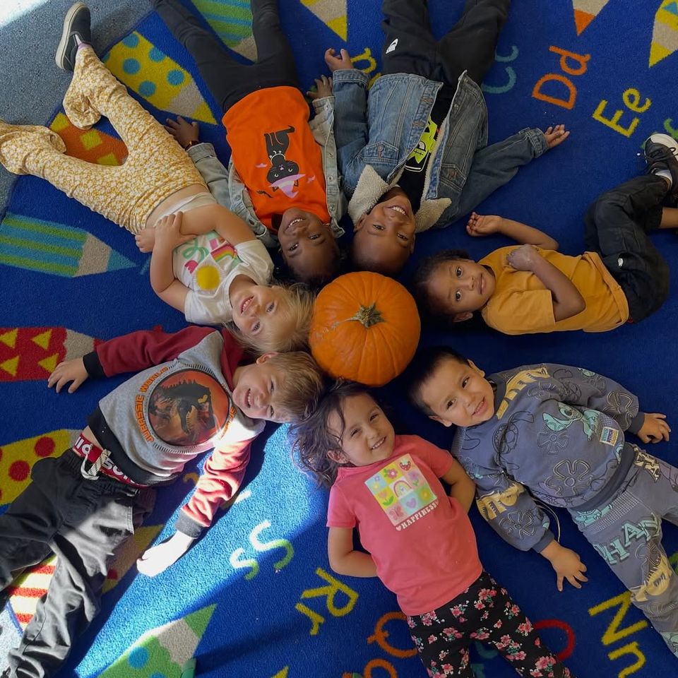 Preschool children lying in a circle around a pumpkin on the alphabet rug during fall fun in Palmdale CA