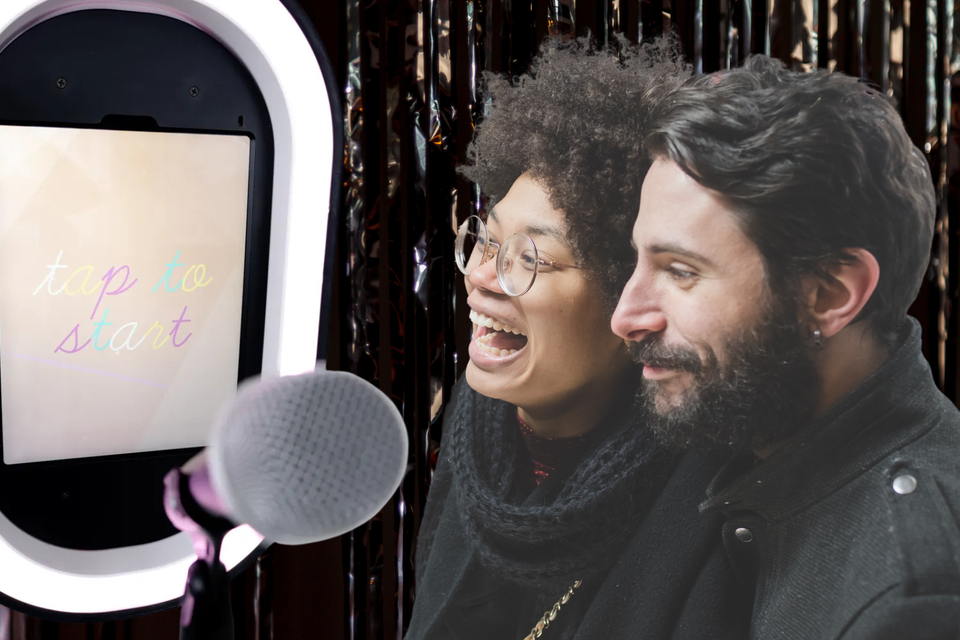 Two guests smiling and laughing while standing in front of a photo booth screen that says tap to start