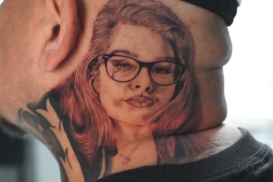 Image of a man in a ball cap with a tattoo of a woman on his neck behind his ear and extend around the back of his head and down to his shoulder in black and reddish ink wearing glasses and wearing a smirk