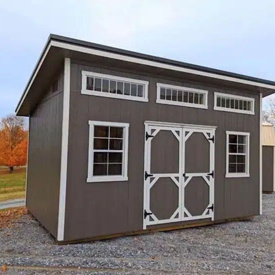 Gray studio shed with transom windows and white double doors on a gravel lot in East Tennessee